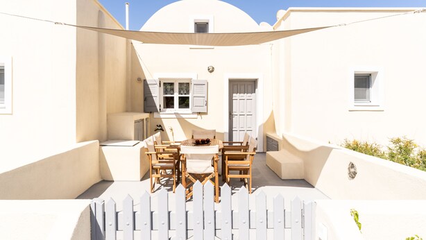 A white-walled building with a patio area featuring a table and chairs, a white picket fence, and a small window.