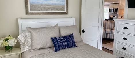 Quarto com cama, mesa de cabeceira com um vaso de flores, quadro com vista para o oceano e porta aberta para uma cozinha.