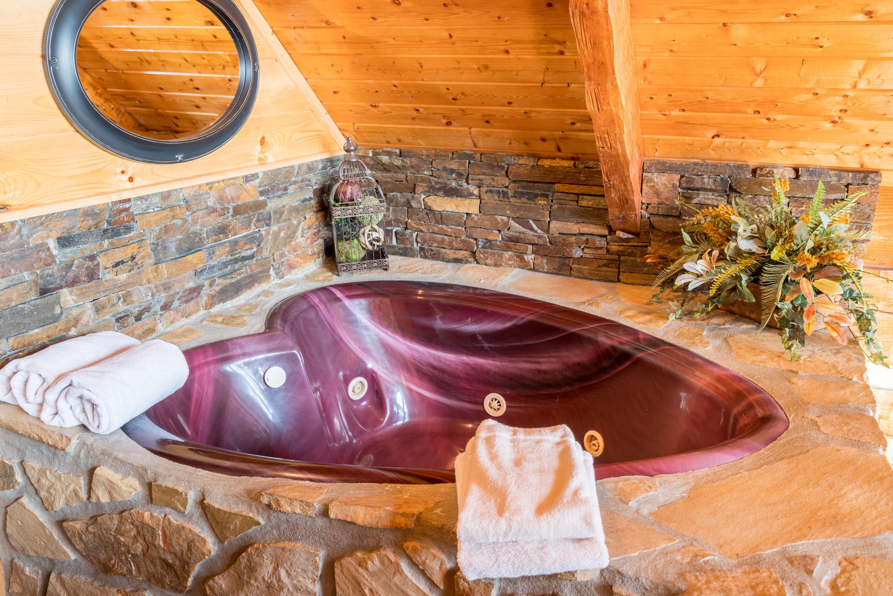 A stone-walled bathroom with a large, oval-shaped bathtub, a wooden ceiling, and a mirror above the tub.