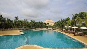 Une grande piscine incurvée, bordée de chaises longues et de parasols, entourée de palmiers et avec un bâtiment en arrière-plan.