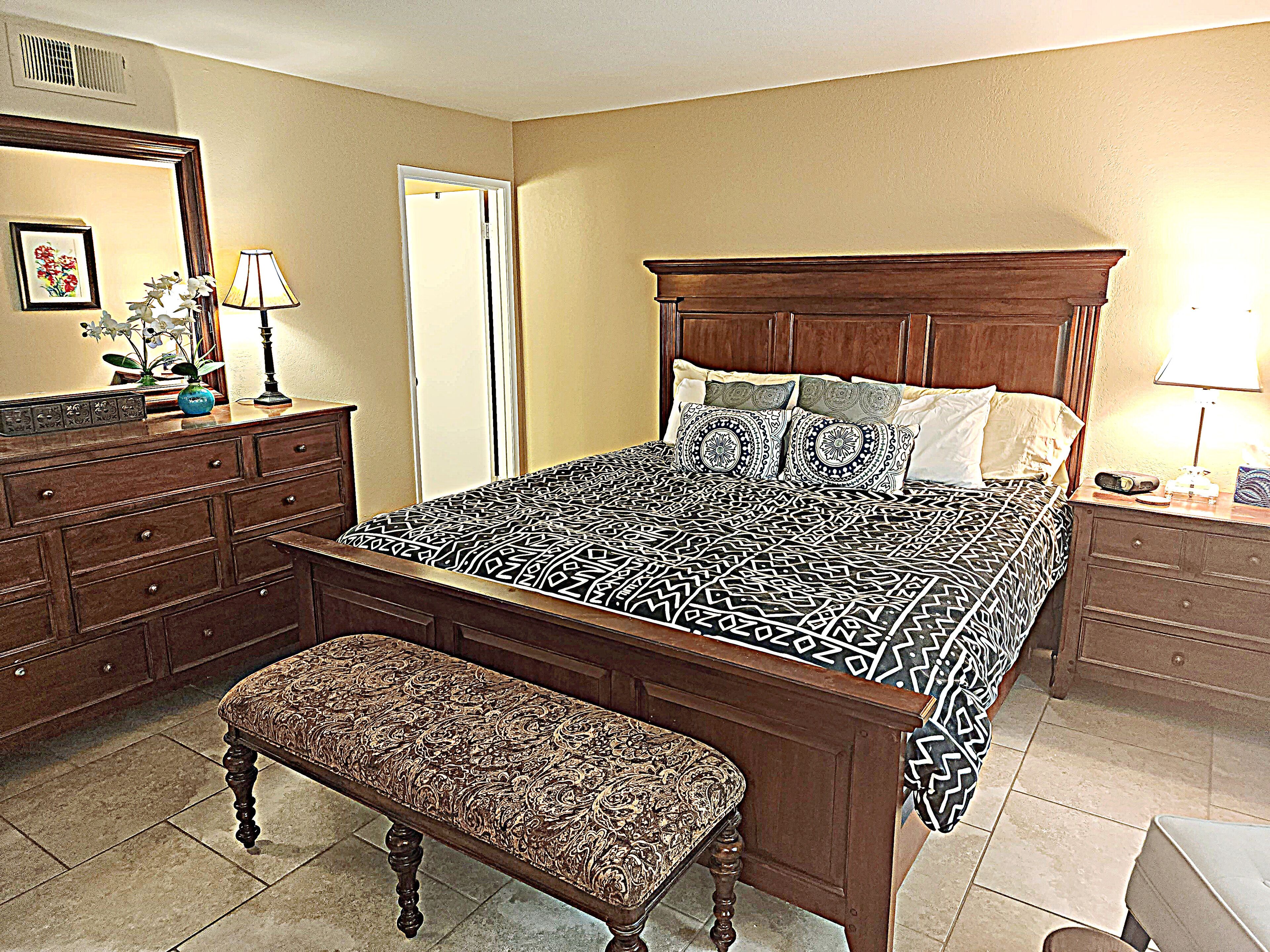 A bedroom with a large bed, a wooden dresser, a bench, and a nightstand with a lamp.