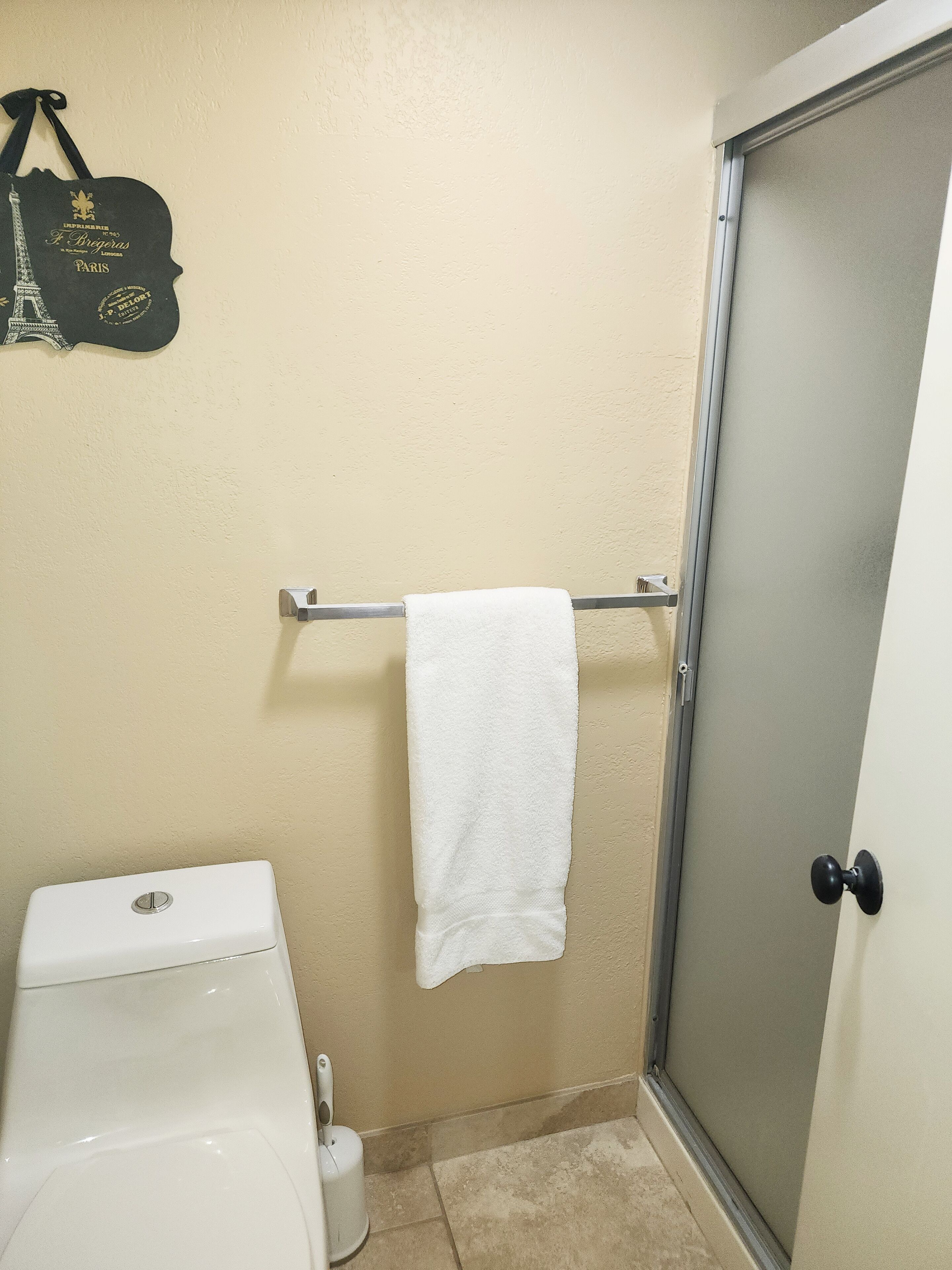 A bathroom with a toilet, a towel rack with a white towel, a black bag with text, and a shower door.