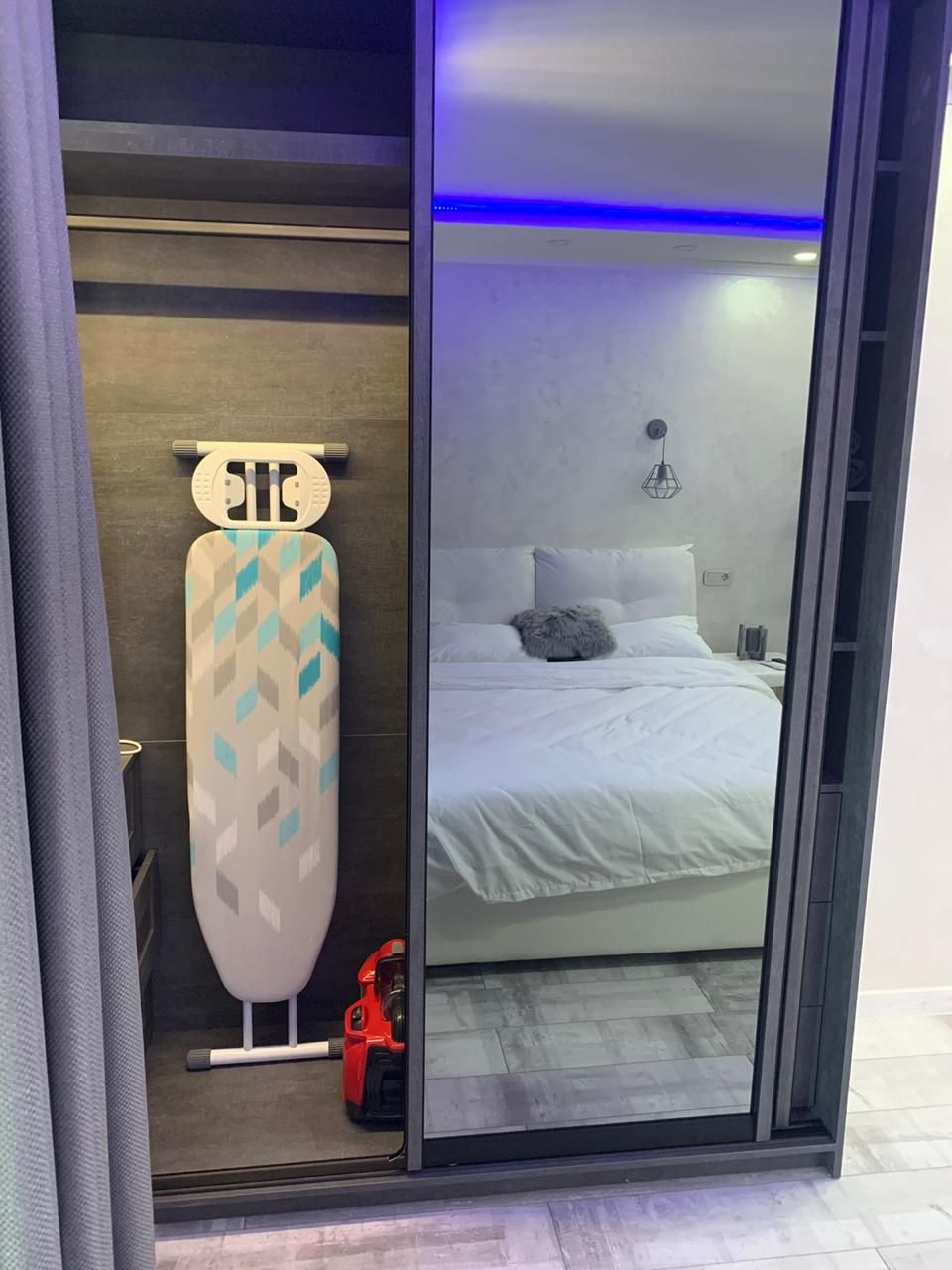 Un dormitorio moderno con cama, tabla de planchar en la pared y una maleta roja.