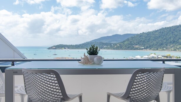 Uma varanda com duas cadeiras brancas, uma mesa de vidro e vista para o mar e montanhas.