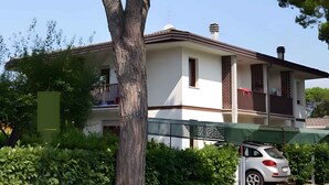 Una casa de dos pisos con un balcón, un coche aparcado en la entrada y un árbol en primer plano.
