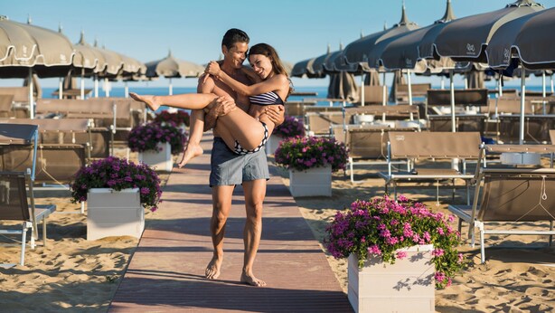 Una pareja se abraza en un paseo marítimo de playa, con sombrillas y tumbonas al fondo.