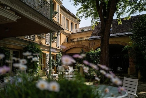A courtyard with a table and chairs, surrounded by greenery and a building with an arched entrance.