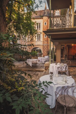 Un espace extérieur aménagé pour les repas, avec des tables et des chaises, entouré de verdure et bordé par un bâtiment doté d’une tour.
