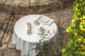 A round table set outdoors with white tablecloth, plates, cups, cutlery, and a teapot.