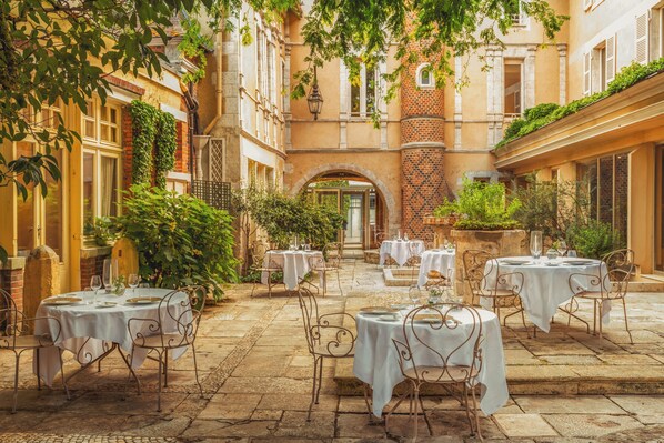 An outdoor dining area with tables and chairs, surrounded by buildings and greenery.