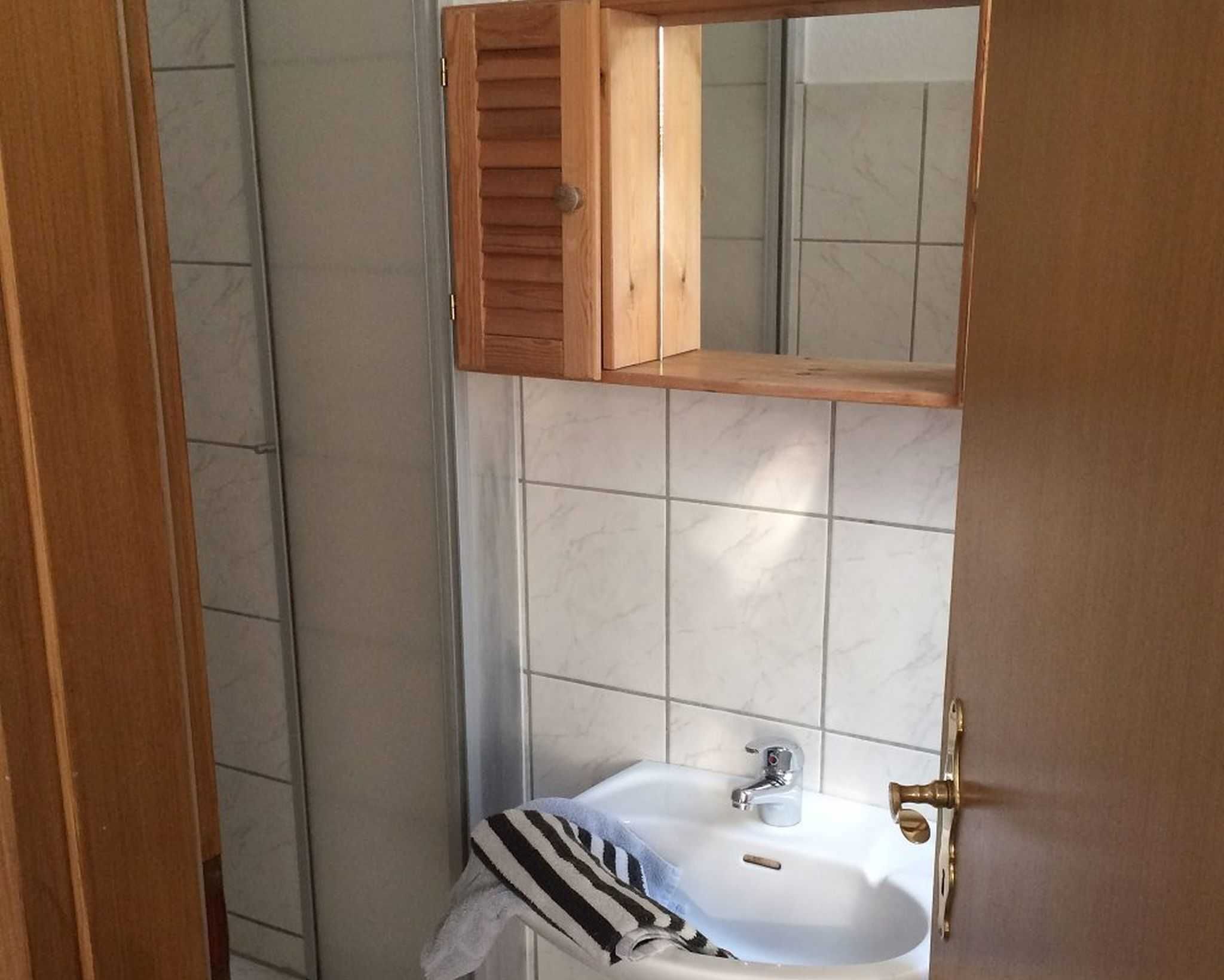 Une salle de bain avec un meuble à pharmacie en bois au-dessus d’un lavabo blanc, sur lequel est accrochée une serviette rayée.