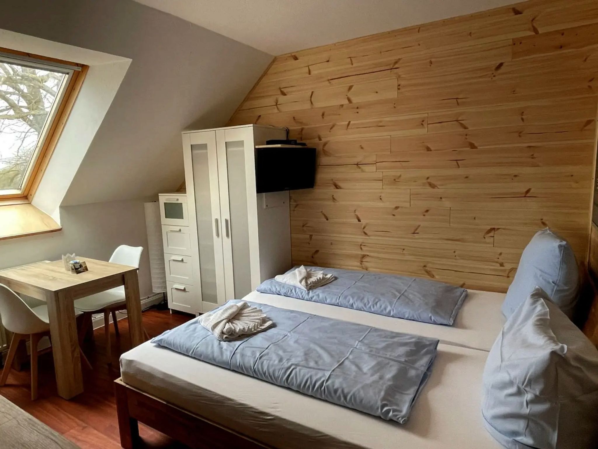 A bedroom with a sloped ceiling, a wooden wall panel, a bed with blue bedding, a desk with a chair, and a wardrobe.