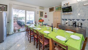 Un coin repas avec une longue table dressée pour six personnes, une cuisine équipée d’une machine à café et un téléviseur fixé au mur.