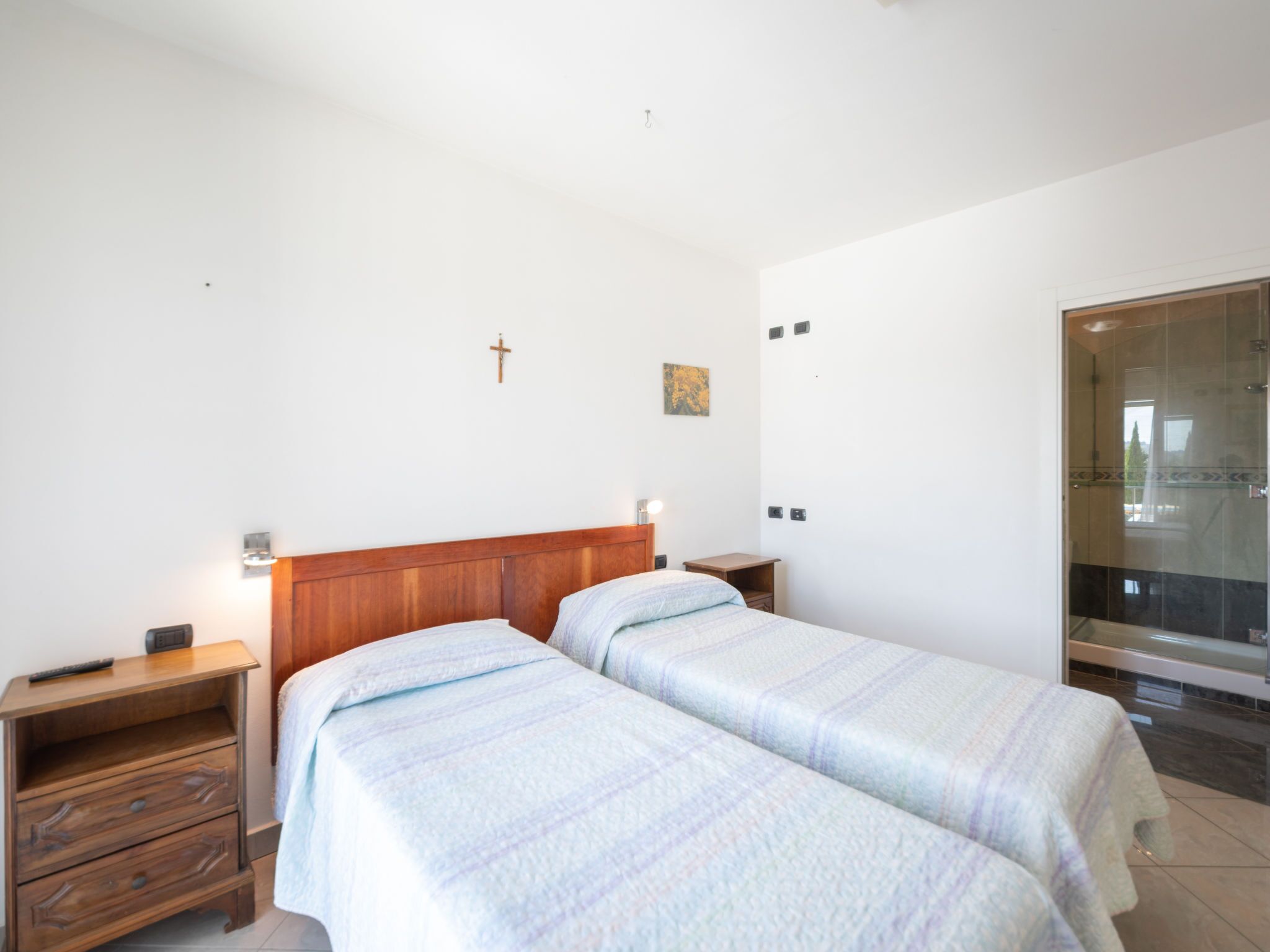 A bedroom with two beds, wooden headboards, bedside tables, a wall-mounted cross, and a glass shower enclosure.