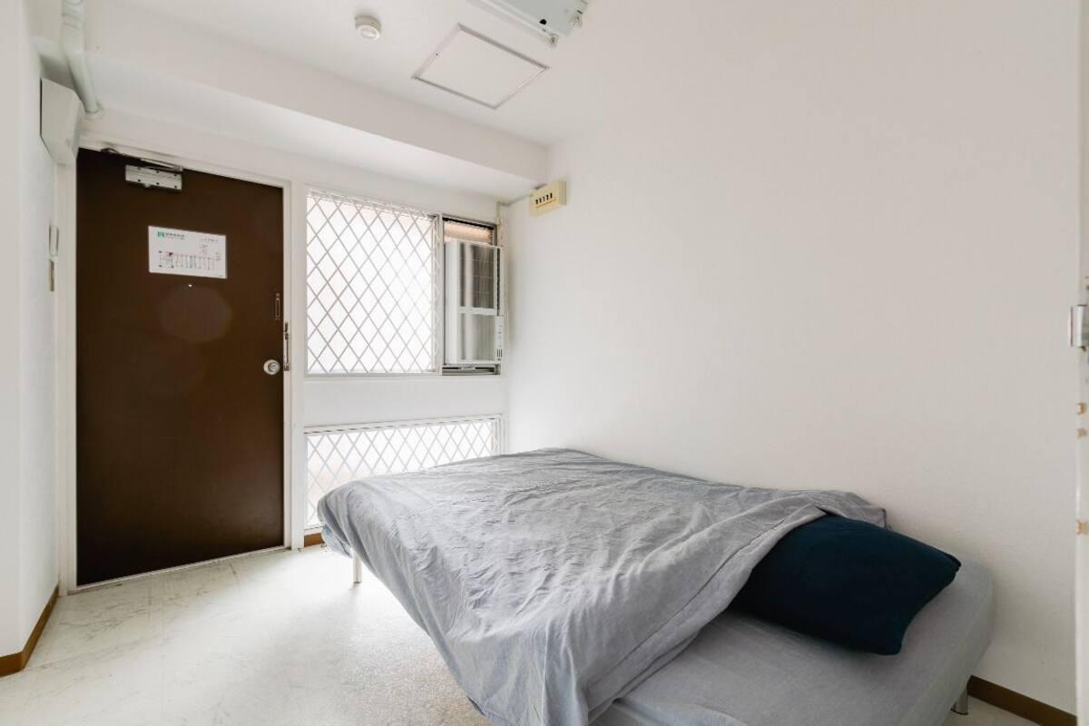 Una habitación pequeña y escasamente amoblada, con una cama individual, una puerta con ventana y un equipo de aire acondicionado a la vista.