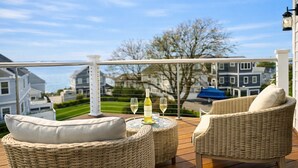 A balcony with wicker furniture, a table with a bottle of wine and glasses, and a view of houses and trees. - Mar Azul \ Walk to the Beach \  Enjoy Ocean Views \  Near Newport (Middletown)