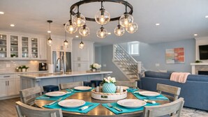 A modern kitchen with white cabinets, a central island, and a dining area with a wooden table set for six. - Mar Azul \ Walk to the Beach \  Enjoy Ocean Views \  Near Newport (Middletown)