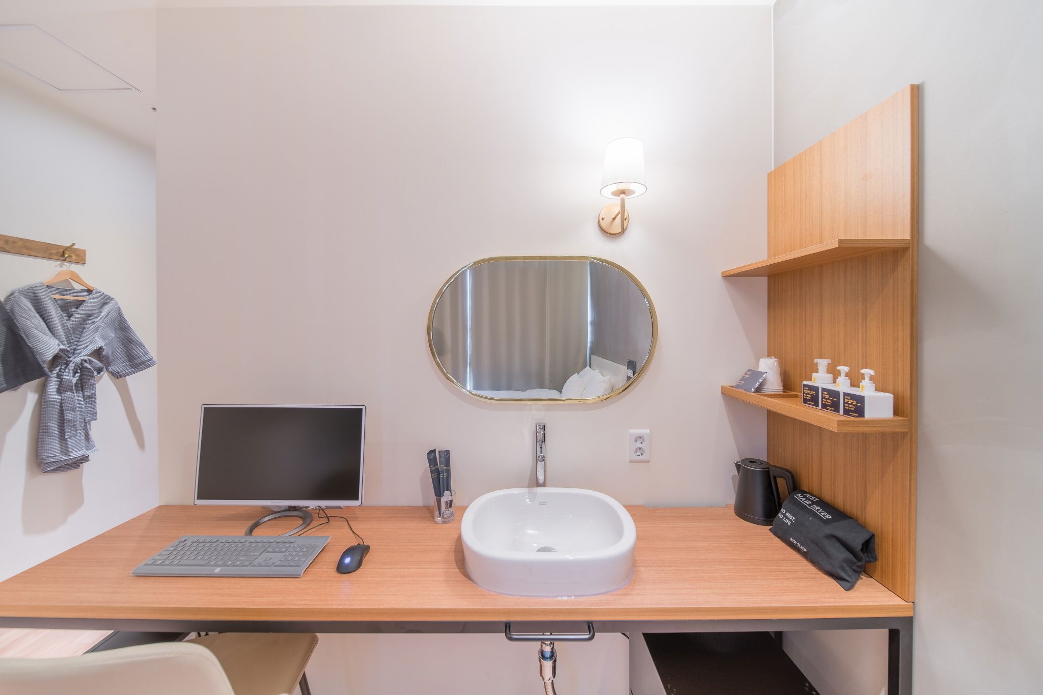 A modern bathroom with a wooden vanity, a round mirror, a sink, a towel rack, a shelf with toiletries, and a desk with a computer and keyboard.