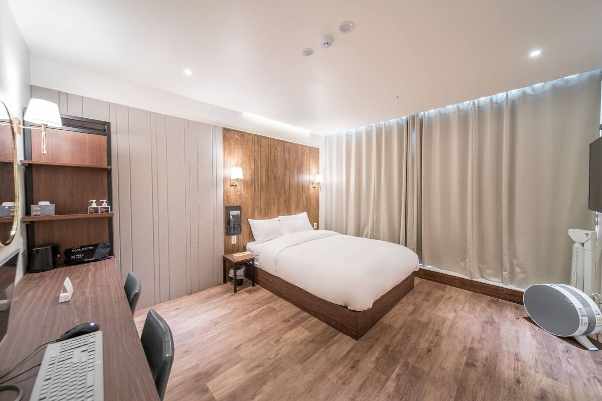 A modern hotel room with a wooden bed, a desk with a computer, a chair, a small table, and a wall-mounted shelf with toiletries.