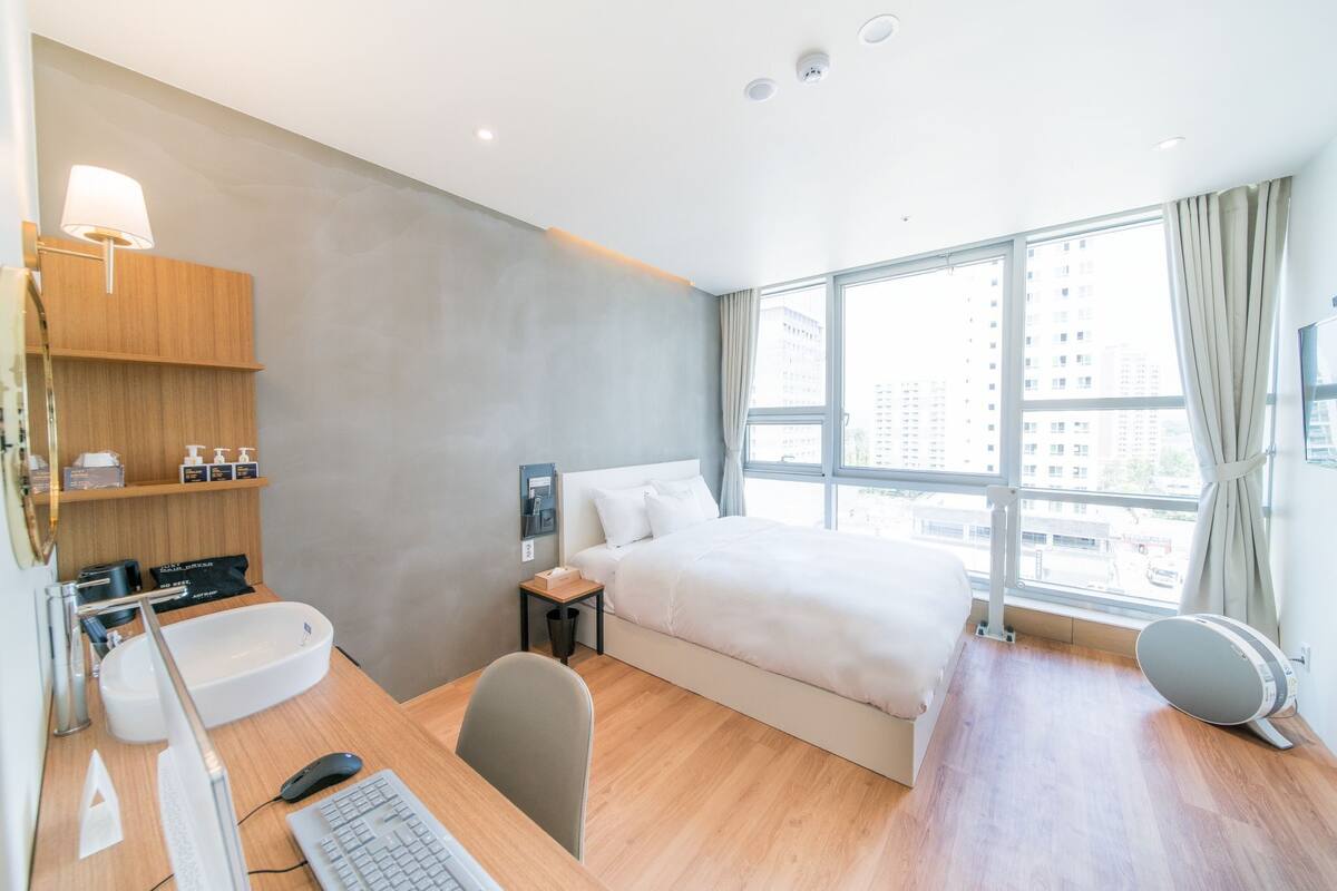 A hotel room with a bed, a desk with a computer, a desk lamp, a small table with a phone, a wall-mounted shelf with toiletries, and a large window with a city view.