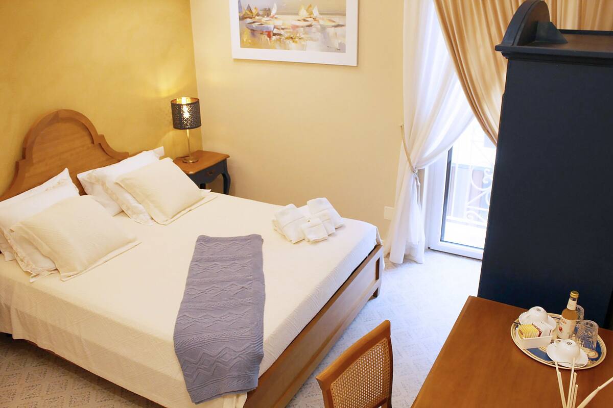 A hotel room with a bed, a nightstand, a lamp, a table with a tray of breakfast items, and a window with curtains.