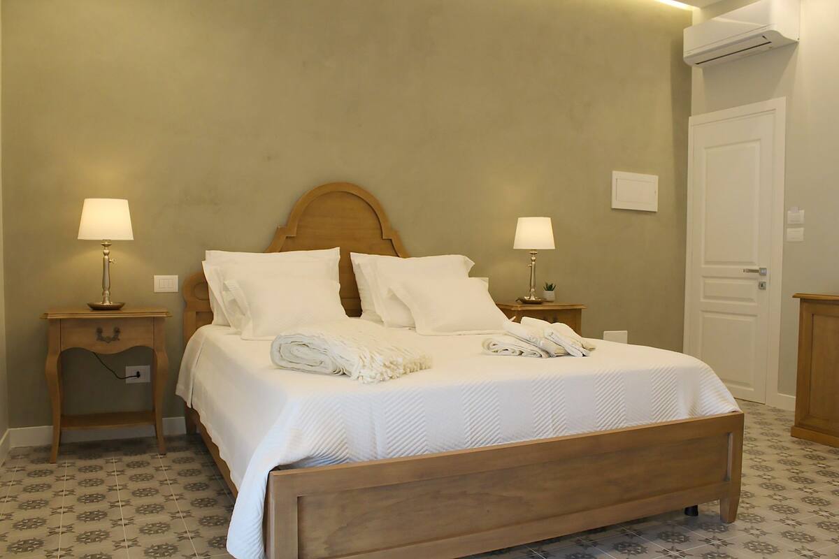 A neatly made bed with white linens and pillows, two bedside tables with lamps, a wooden headboard, and patterned floor tiles.