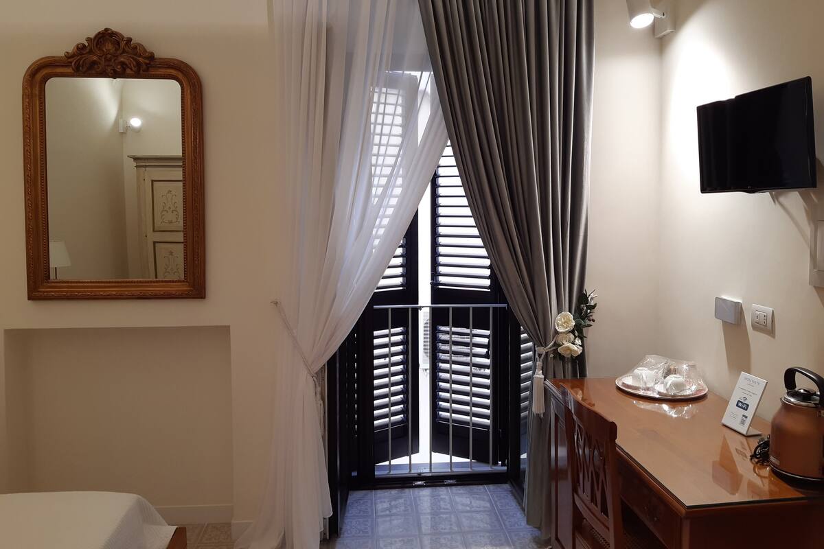 A hotel room with a large mirror, a wooden dresser, a flat-screen TV, and a view of the outside through a window with blinds.