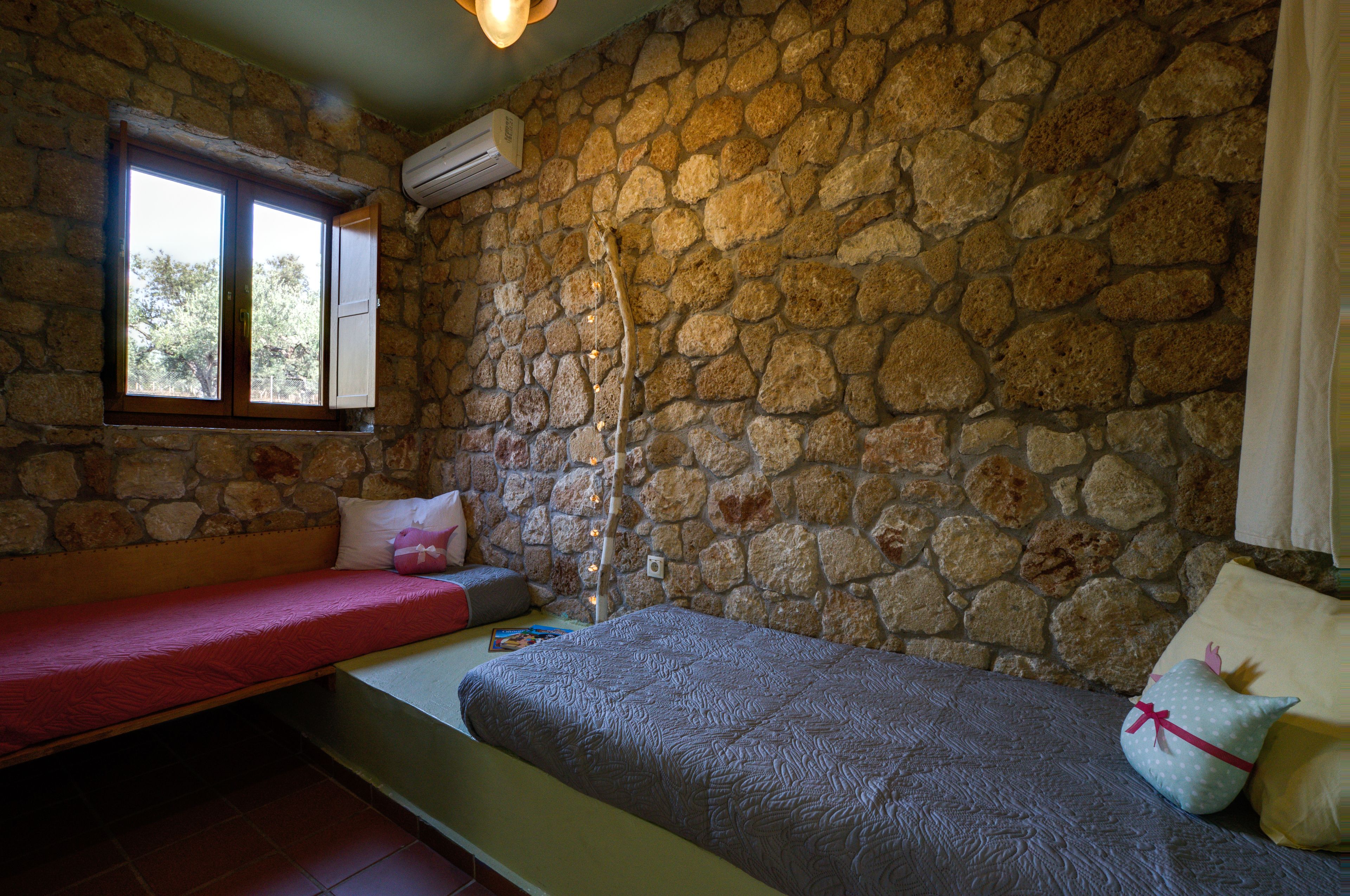 Ein Zimmer mit einer Steinsandsteinwand, einem Fenster mit Holzrahmen, einem Bett mit grauer Tagesdecke und einer Bank mit roter Auflage.