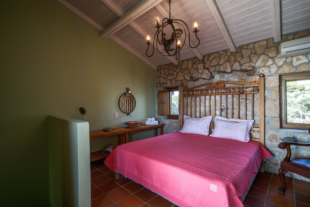 A bedroom with a stone wall, a wooden bed frame, a chandelier, and a window with a view of trees.