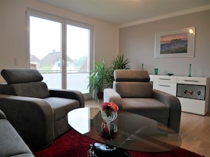 A modern living room with two grey sofas, a glass coffee table, a red rug, a potted plant, and a white cabinet with decorative items.