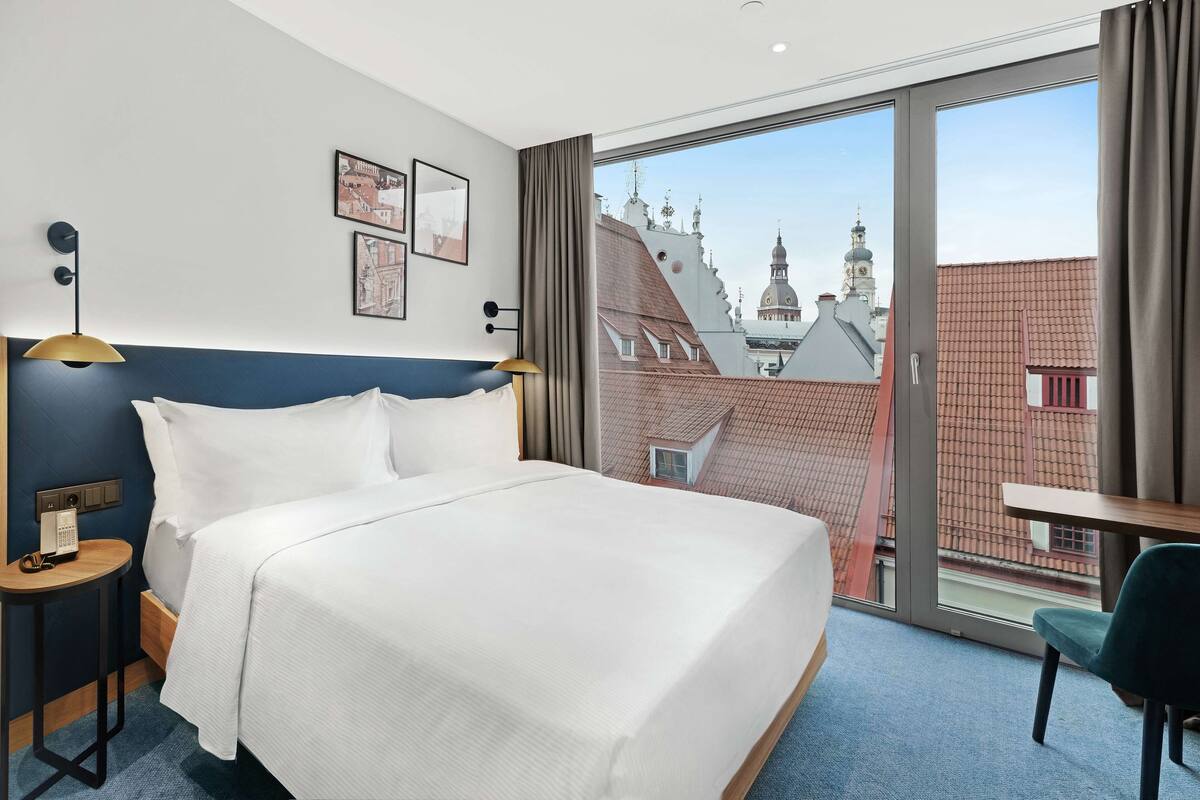 Hotellihuone, jossa on suuri sänky, työpöytä, tuoli, lamppu ja suuri ikkuna, josta on näkymä ympäröiviin rakennuksiin.