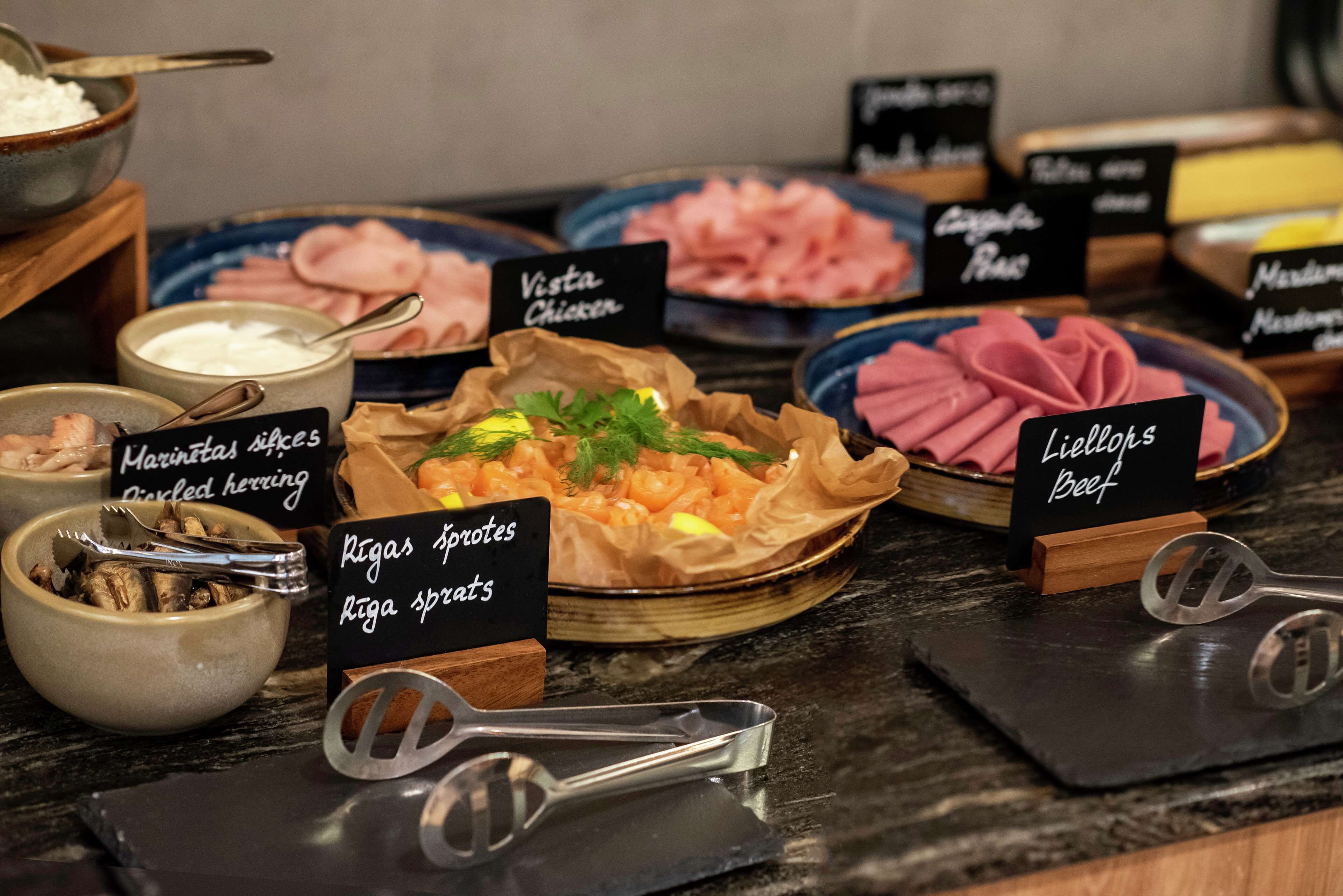 Un bufé con diversos platos etiquetados, incluyendo 'Marinatas', 'Riga sprutes', 'Vesta Chicken', 'Lihlohs Beef' y 'Capgras'.