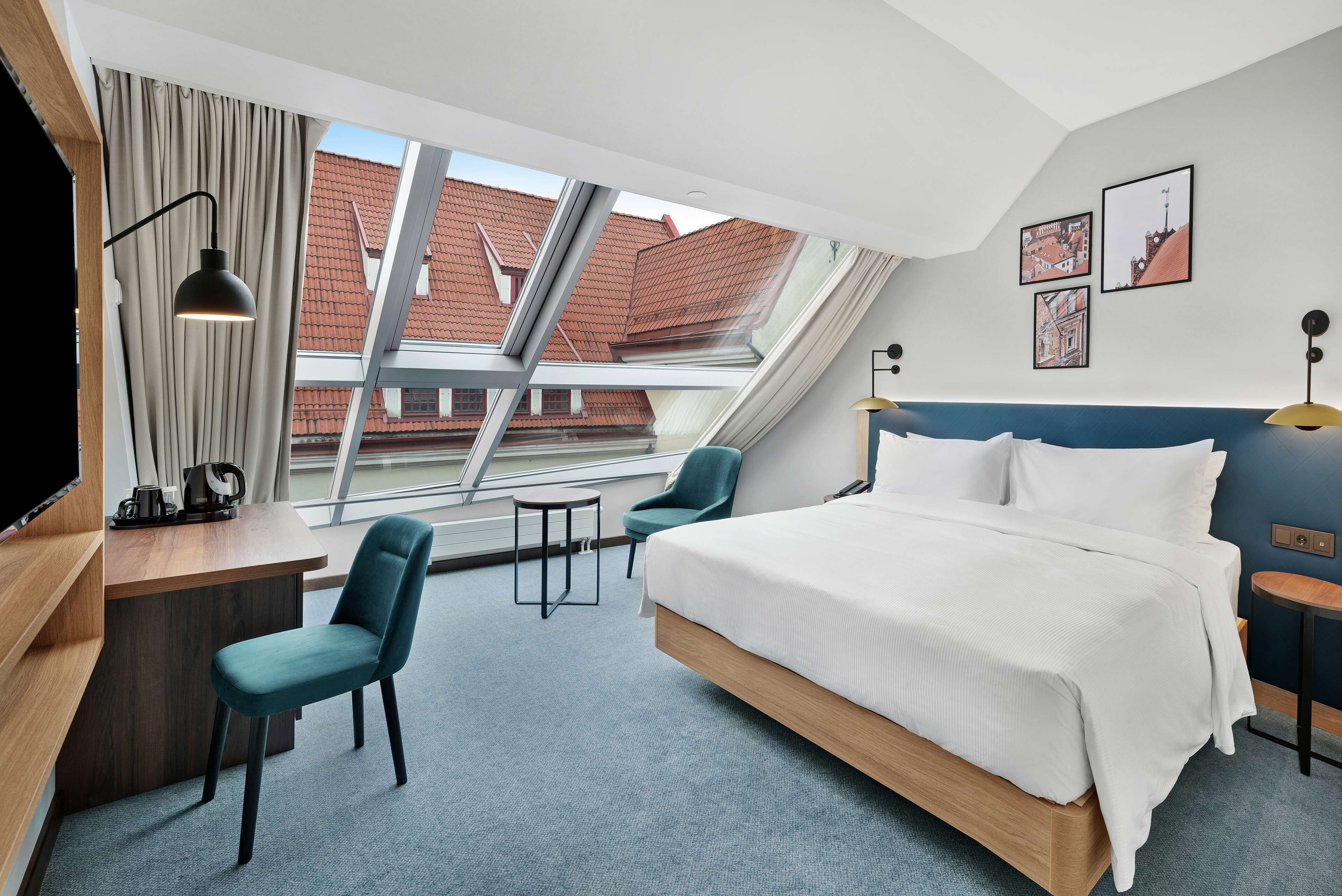 A modern hotel room with a large bed, a desk, a chair, and a view of the city through a skylight.