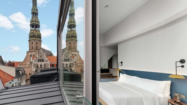Habitación de hotel con cama, mesita de noche y vistas a edificios históricos con torres.