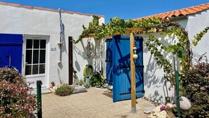 A white building with a blue door and a blue shutter, surrounded by greenery and a clear sky. - La Cabane de pêche de la Cotinière (Saint-Pierre-d´Oléron)