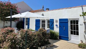 A white building with blue shutters and a white door, a satellite dish, and a small garden area with pink flowers. - La Cabane de pêche de la Cotinière (Saint-Pierre-d´Oléron)