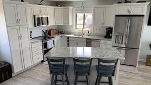 A modern kitchen with white cabinets, a stainless steel refrigerator, a built-in oven, and a marble countertop island with bar stools. - Sounds Groovy OBX: Waterfront Home with Dock (Nags Head)