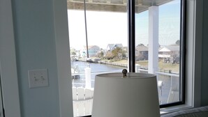 A room with a window overlooking a water body and buildings. There is a lamp on a table in front of the window. - Sounds Groovy OBX: Waterfront Home with Dock (Nags Head)