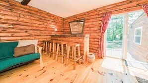 A log cabin interior with a wooden floor, a green sofa, wooden stools, a mirror, and a window with curtains. - Super Clean, Pet Friendly Cottage! 3 Bedroom! NO EXTRA FEES!! (Alexandria Bay)