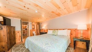 A bedroom with a wooden ceiling, a large bed with patterned bedding, a nightstand with a lamp, and a wardrobe. - Super Clean, Pet Friendly Cottage! 3 Bedroom! NO EXTRA FEES!! (Alexandria Bay)