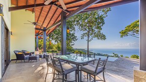 A covered outdoor dining area with a glass table and chairs, overlooking a scenic view of the sea.