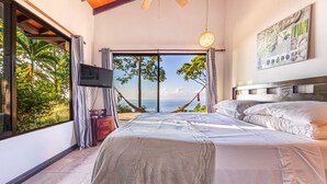 A bedroom with a bed, a TV, a hammock, and a view of the ocean.