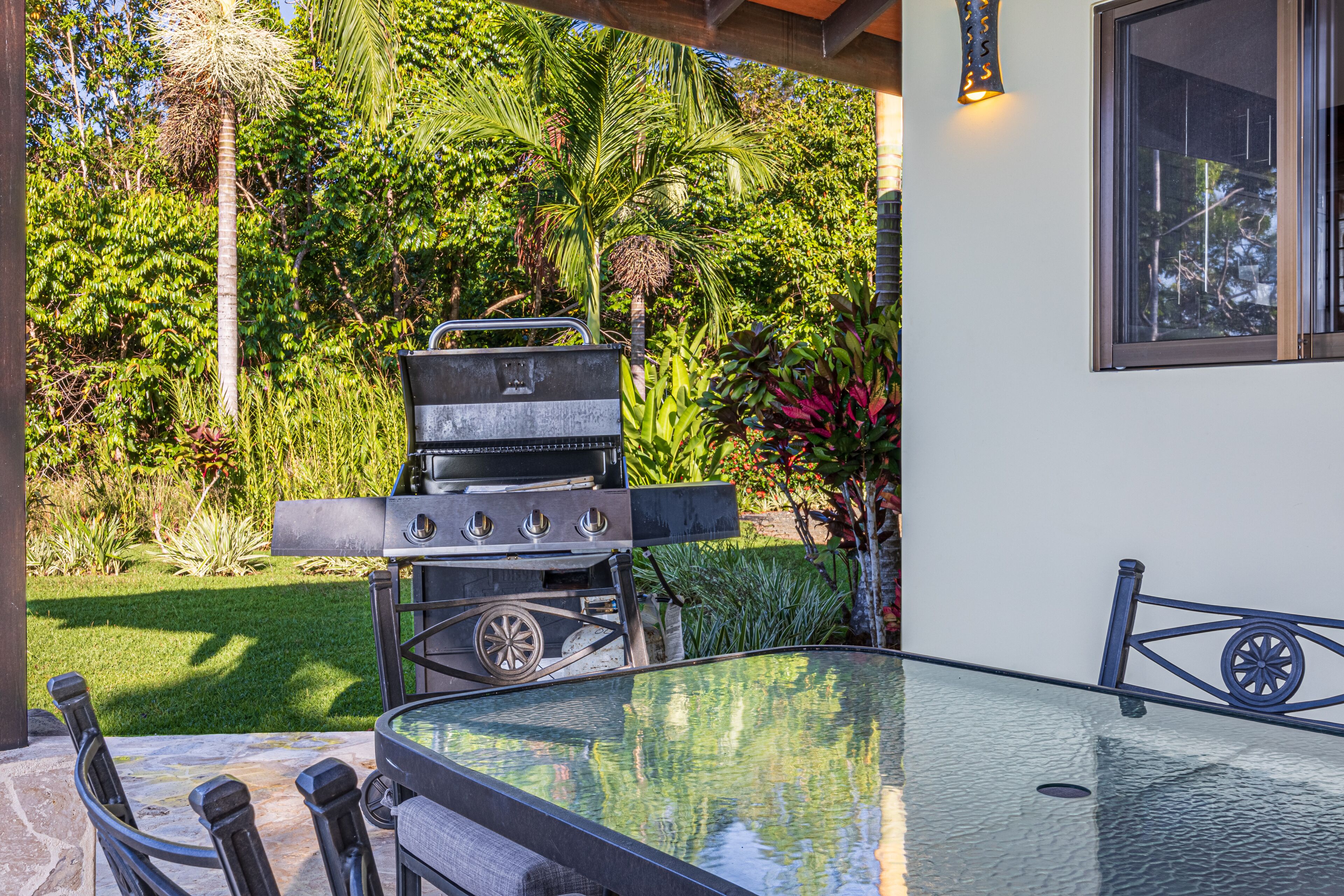 Eine Terrassenanlage im Freien mit einem Glastisch, Metallstühlen, einem Grill und üppiger Bepflanzung.