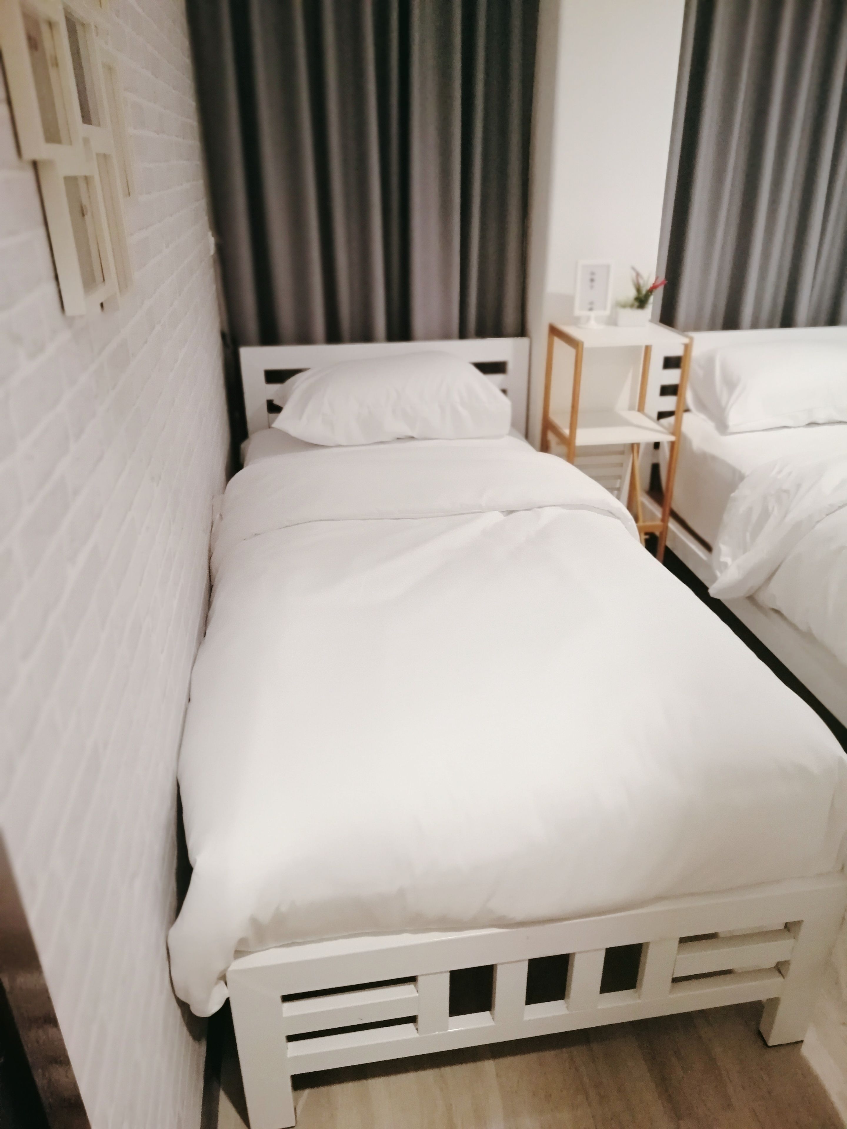 A single white bed with a wooden headboard in a room with white brick walls and a grey curtain.