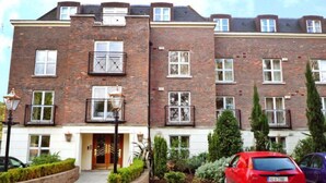 A multi-story brick apartment building with a central entrance, a small garden area, and a red car parked in front.