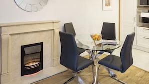 A dining area with a glass table, four chairs, a fireplace, and a kitchen in the background.