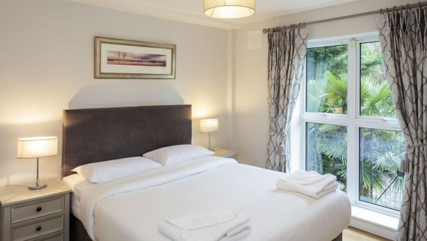 A neatly made bed with white linens, two bedside lamps, a framed picture on the wall, and a window with curtains.