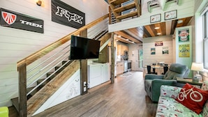 Ein moderner Loft-Wohnraum mit freiliegenden Holzbalken, einer Treppe, einem Flachbildfernseher, einer Küchenzeile und einer gemütlichen Sitzecke mit einem Kissen mit Muster.