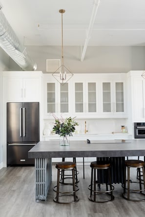 A modern kitchen with white cabinets, a stainless steel refrigerator, a central island with stools, and a pendant light. - 2 Luxury Suites - Full Kitchen, 1 Kitchenette, 2 King Bed, 1 Full, 1 Twin,2 Bath (Logan)
