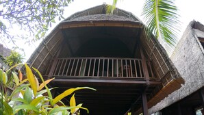 Une construction à toit de chaume, dotée d’un balcon en bois et entourée de verdure.